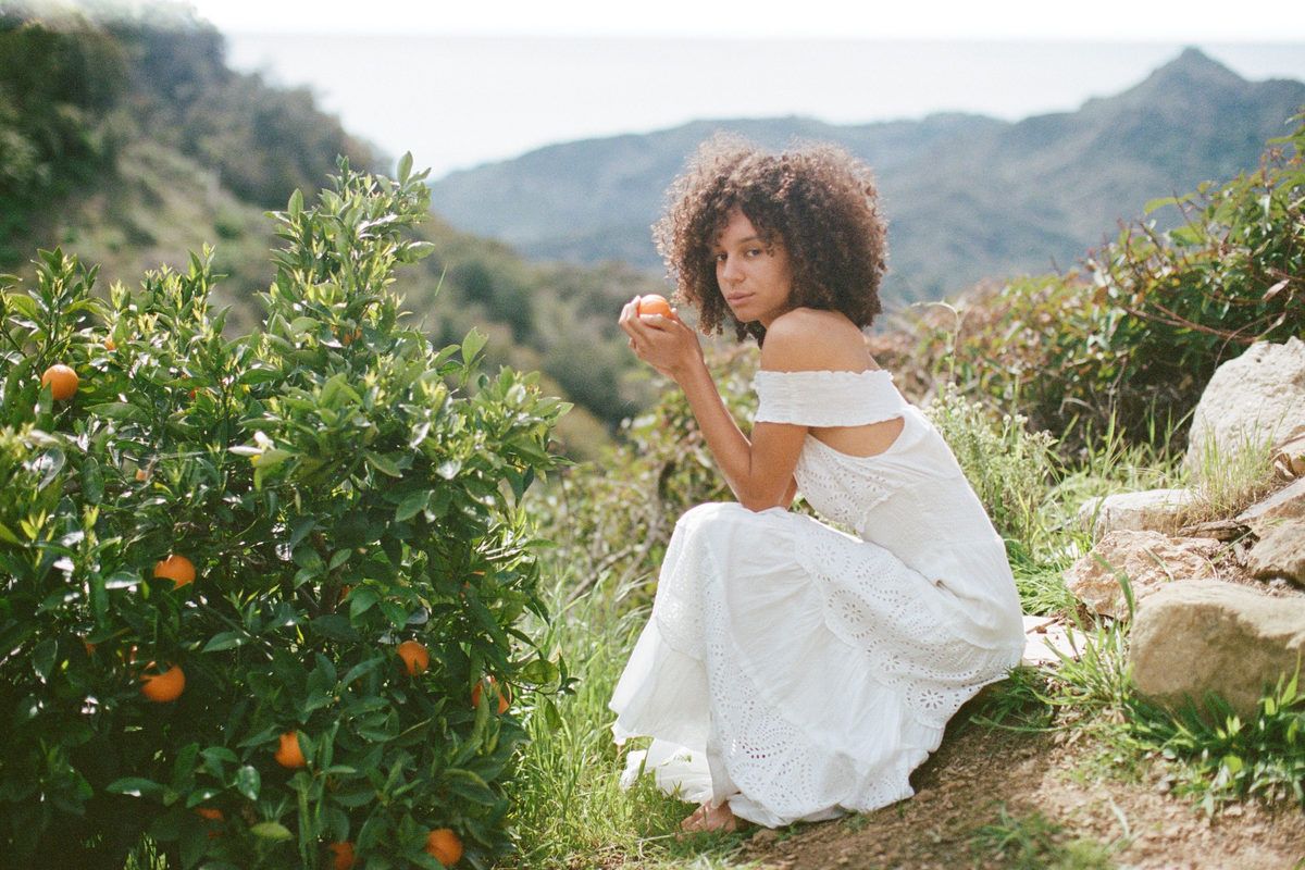 A woman crouching beside a citrus tree on a hillside in soft daylight.