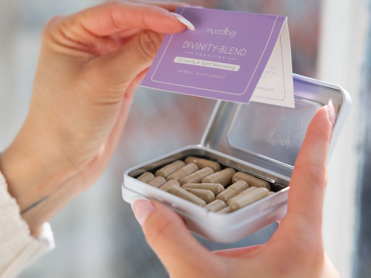 A pair of hands opening a tin of Mycrologi Divinity Blend capsules beside a window.