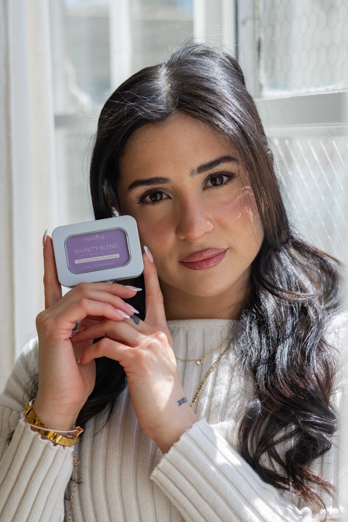 A woman holding a tin of Mycrologi capsules beside her face in warm daylight.