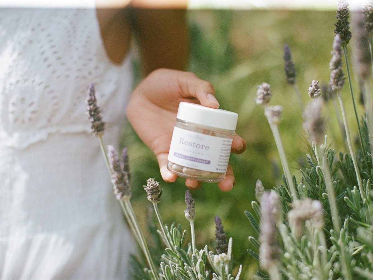 A hand holding a jar of Mycrologi capsules among lavender in a sunlit garden.