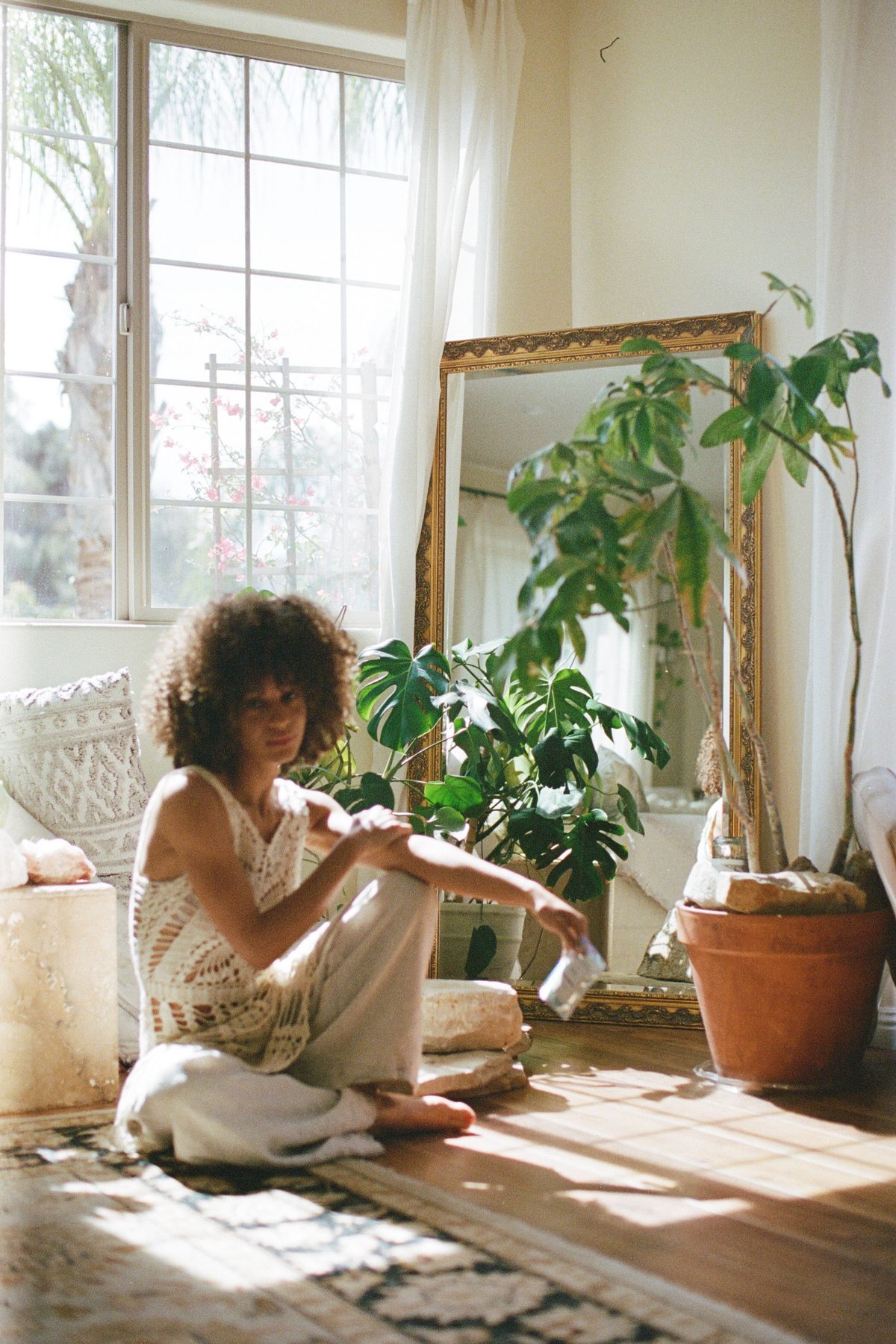 A person sitting cross legged on the floor of a plant filled room in afternoon light.